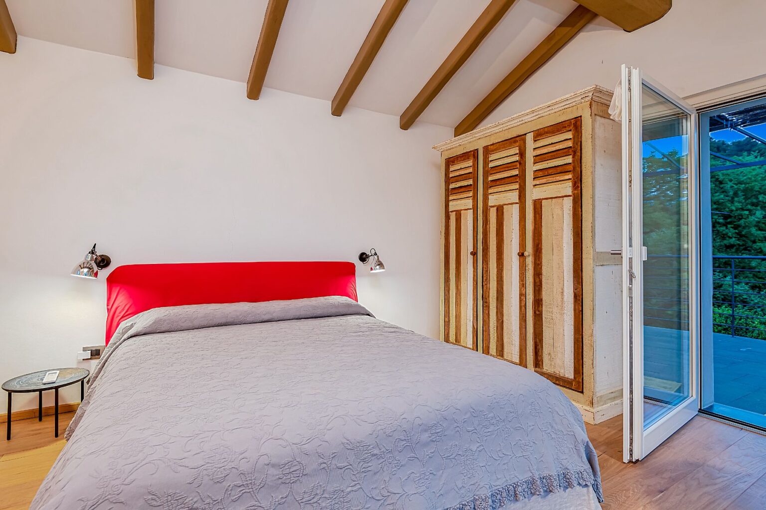 Cozy bedroom featuring a red upholstered headboard, gray bedding, and a wooden wardrobe. The room has wooden beams on the ceiling and a sliding glass door that opens to a balcony, providing natural light and a view of greenery. Perfect for a relaxing getaway. | Only on SopranoVillas