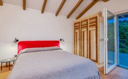 Cozy bedroom featuring a red upholstered headboard, gray bedding, and a wooden wardrobe. The room has wooden beams on the ceiling and a sliding glass door that opens to a balcony, providing natural light and a view of greenery. Perfect for a relaxing getaway.