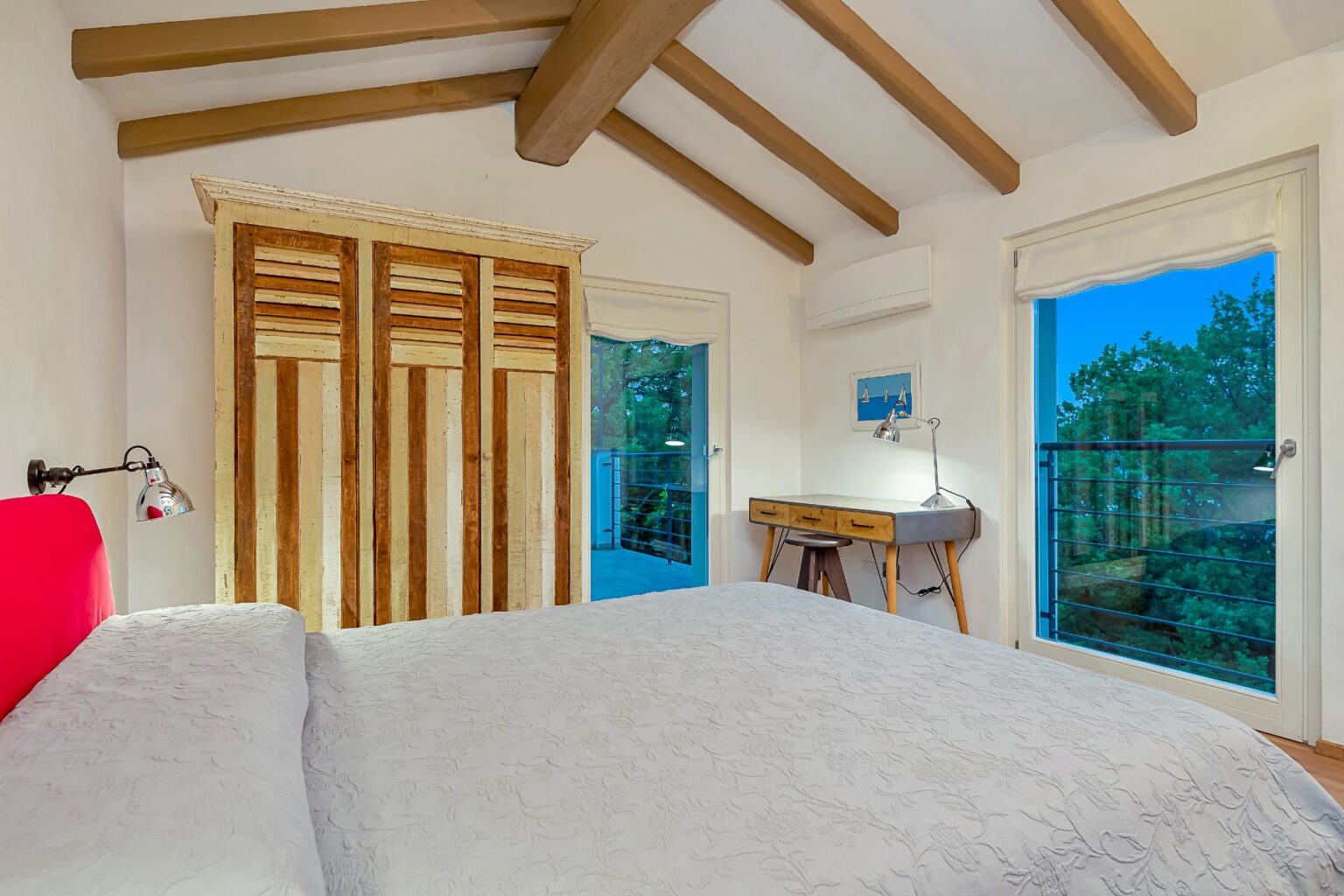 Cozy bedroom featuring a king-sized bed with a light gray quilt, wooden ceiling beams, and a vintage wardrobe. Natural light flows through large windows, showcasing a balcony view surrounded by greenery. A small wooden desk with a stylish lamp complements the space, creating an inviting atmosphere. Ideal for relaxation and comfort. | Only on SopranoVillas