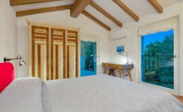 Cozy bedroom featuring a king-sized bed with a light gray quilt, wooden ceiling beams, and a vintage wardrobe. Natural light flows through large windows, showcasing a balcony view surrounded by greenery. A small wooden desk with a stylish lamp complements the space, creating an inviting atmosphere. Ideal for relaxation and comfort.