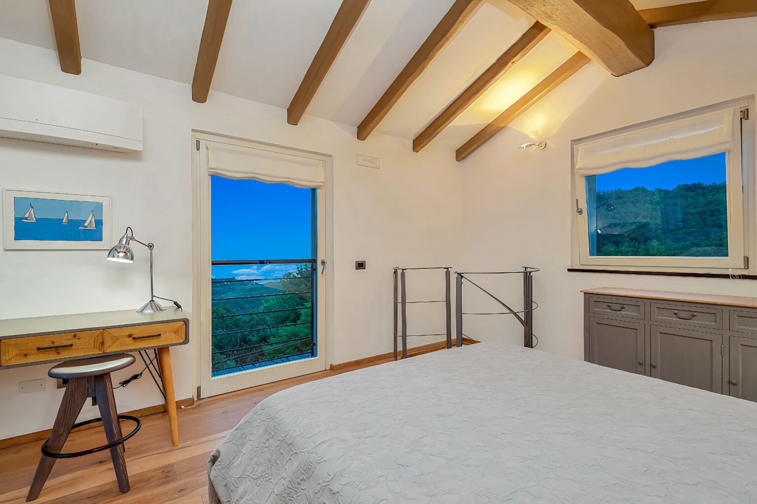 Modern bedroom with wooden beams, a cozy bed, and a desk featuring a lamp. Large window offers a scenic view of greenery and blue sky. Natural light brightens the space, enhancing the minimalist decor. | Only on SopranoVillas