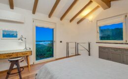 Modern bedroom with wooden beams, a cozy bed, and a desk featuring a lamp. Large window offers a scenic view of greenery and blue sky. Natural light brightens the space, enhancing the minimalist decor.