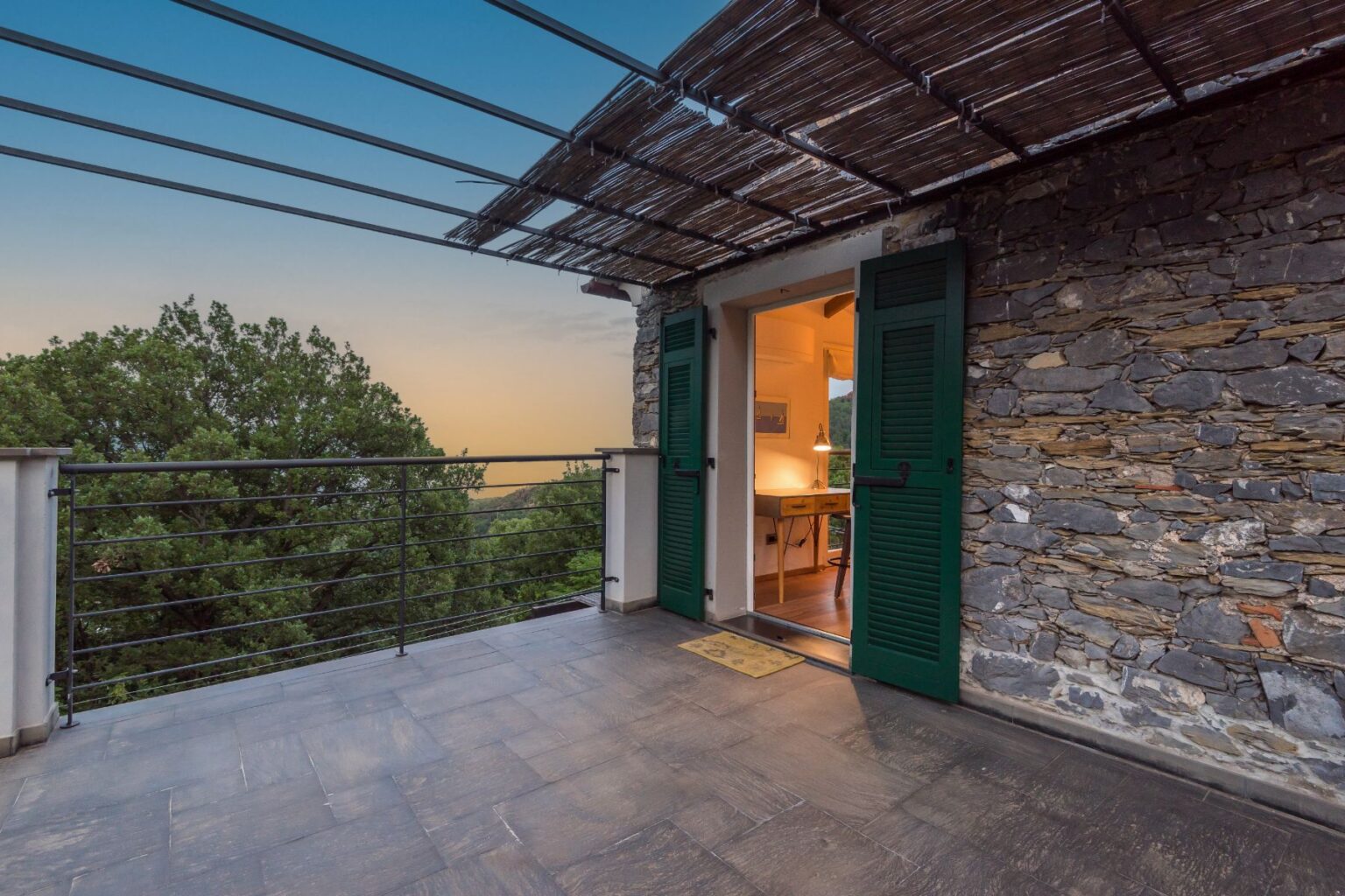 Modern balcony with green shutters overlooking lush greenery, showcasing a cozy interior with a desk and warm lighting, set against a sunset sky. | Only on SopranoVillas