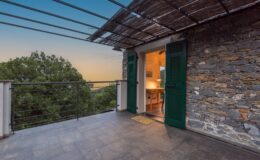 Modern balcony with green shutters overlooking lush greenery, showcasing a cozy interior with a desk and warm lighting, set against a sunset sky.