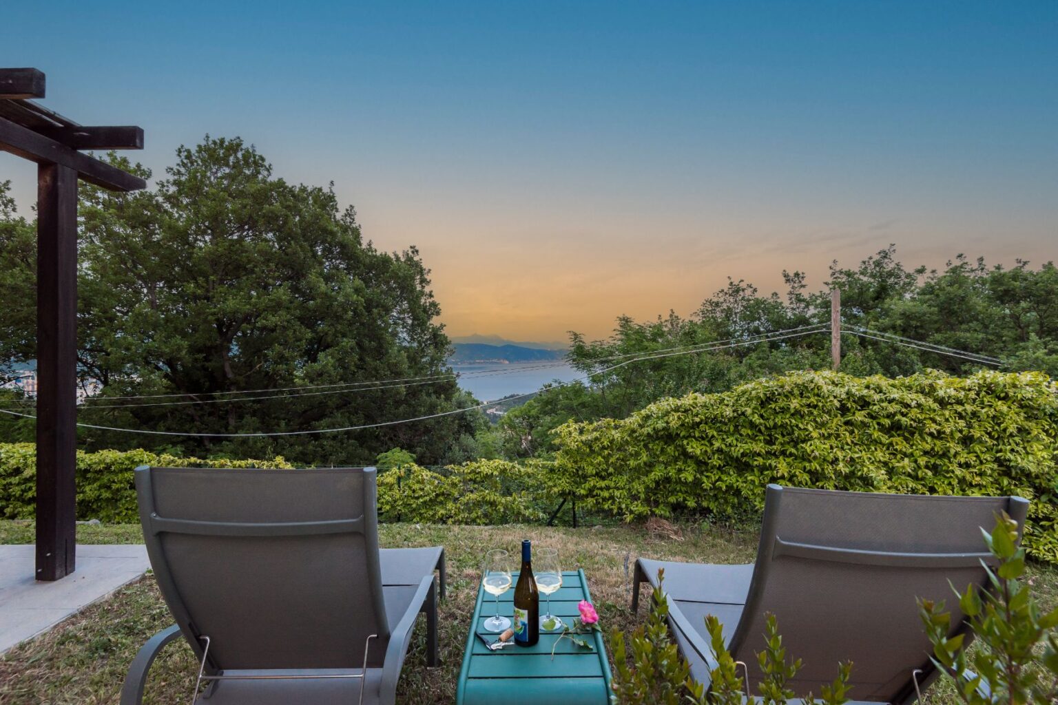Relaxing outdoor seating area with two lounge chairs overlooking a scenic view of the ocean and mountains at sunset, featuring a table with wine glasses and a bottle on a green table, surrounded by lush greenery. | Only on SopranoVillas
