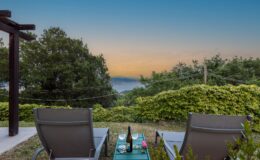 Relaxing outdoor seating area with two lounge chairs overlooking a scenic view of the ocean and mountains at sunset, featuring a table with wine glasses and a bottle on a green table, surrounded by lush greenery.