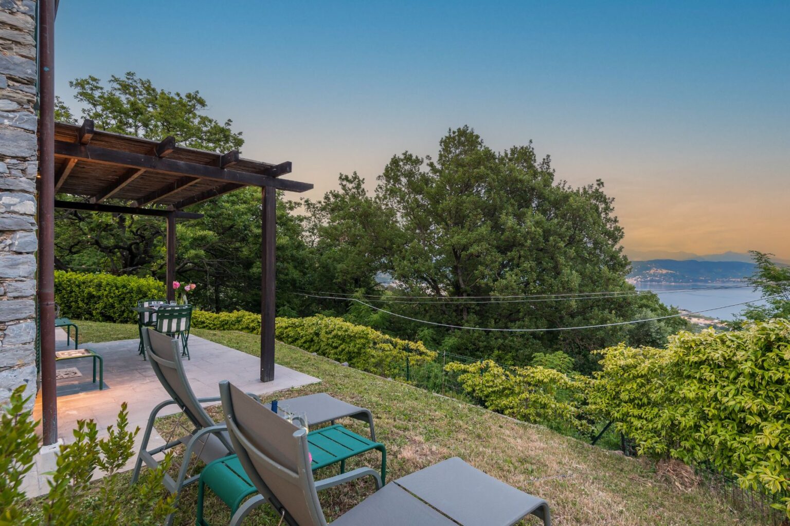 Scenic outdoor patio with lounge chairs and a small table, surrounded by lush greenery and overlooking a tranquil landscape at sunset. Ideal for relaxation and enjoying nature. | Only on SopranoVillas