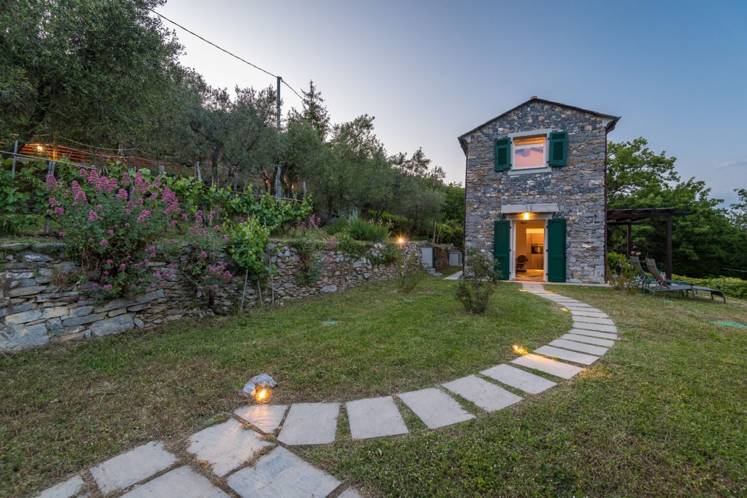 Charming stone house with green shutters surrounded by lush gardens and flowering plants, featuring a winding stone pathway leading to the entrance, set against a twilight sky. Ideal for a serene getaway in nature. | Only on SopranoVillas