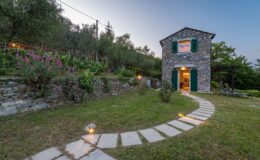 Charming stone house with green shutters surrounded by lush gardens and flowering plants, featuring a winding stone pathway leading to the entrance, set against a twilight sky. Ideal for a serene getaway in nature.