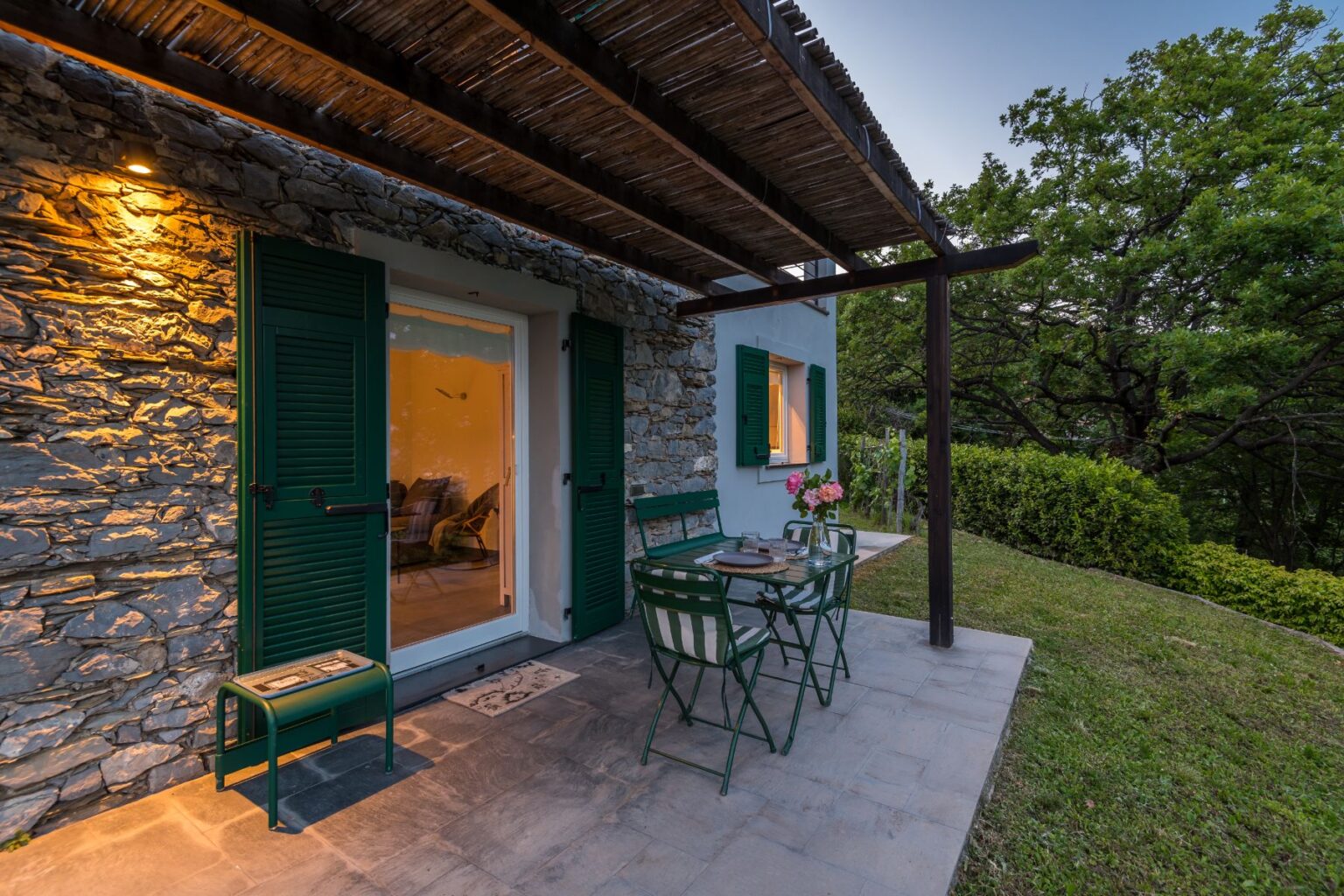 Charming outdoor patio of a stone cottage featuring green shutters, a dining table with chairs, and decorative flowers, surrounded by lush greenery and soft evening lighting. Ideal for relaxing and enjoying nature. | Only on SopranoVillas