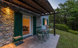 Charming outdoor patio of a stone cottage featuring green shutters, a dining table with chairs, and decorative flowers, surrounded by lush greenery and soft evening lighting. Ideal for relaxing and enjoying nature.