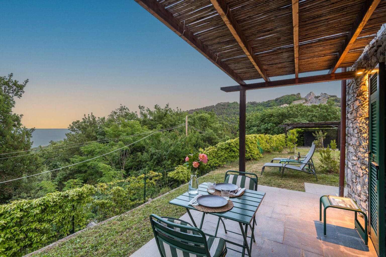 Cozy outdoor patio with a table set for dining, featuring green chairs and a vase of flowers, surrounded by lush greenery and a view of the sea at sunset. Perfect for relaxation and outdoor meals in a tranquil setting. | Only on SopranoVillas