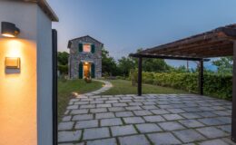 Stone cottage with green shutters surrounded by landscaped garden and pathway, featuring a wooden pergola, captured during twilight.