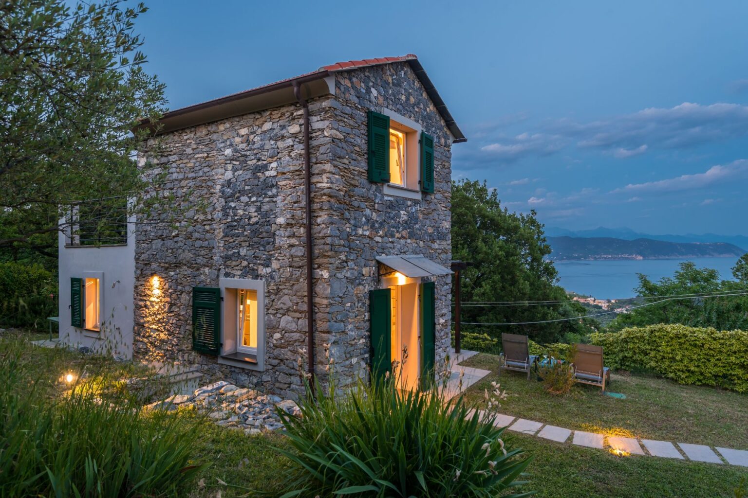 Charming stone cottage with green shutters, set against a twilight sky, featuring warm lights glowing from the windows. Surrounded by lush greenery and a scenic view of the coastline, the outdoor area includes two lounge chairs and a stone pathway leading to the entrance. Ideal for a serene getaway. | Only on SopranoVillas