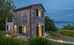 Charming stone cottage with green shutters, set against a twilight sky, featuring warm lights glowing from the windows. Surrounded by lush greenery and a scenic view of the coastline, the outdoor area includes two lounge chairs and a stone pathway leading to the entrance. Ideal for a serene getaway.