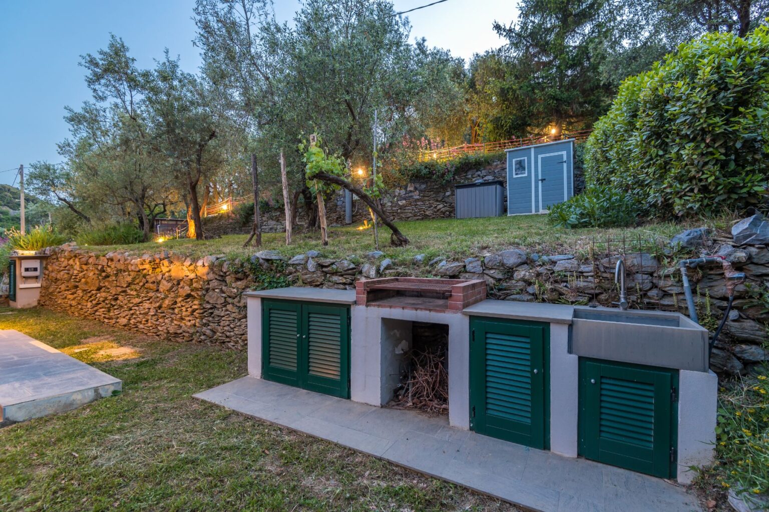 Outdoor kitchen area featuring stone walls, green shuttered storage units, and a brick grill, surrounded by lush greenery and illuminated pathways. Ideal for entertaining in a garden setting. | Only on SopranoVillas