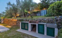 Outdoor kitchen area featuring stone walls, green shuttered storage units, and a brick grill, surrounded by lush greenery and illuminated pathways. Ideal for entertaining in a garden setting.