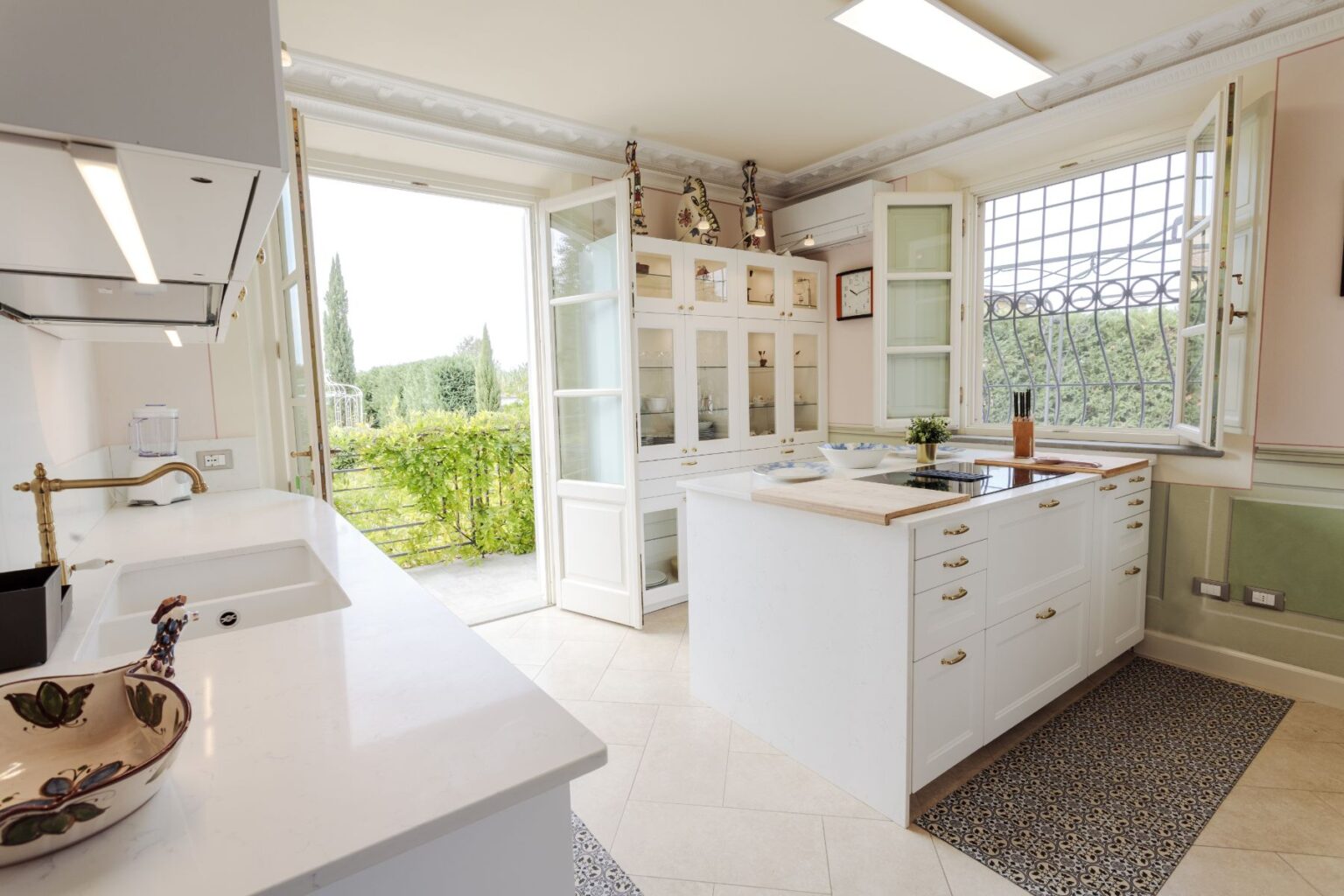 Bright and airy modern kitchen featuring white cabinetry, a central island with a wooden countertop, and large windows opening to a scenic outdoor view. The space includes a stylish sink, a blend of decorative elements, and a cozy atmosphere enhanced by natural light. Ideal for home cooking and entertaining. | Only on SopranoVillas