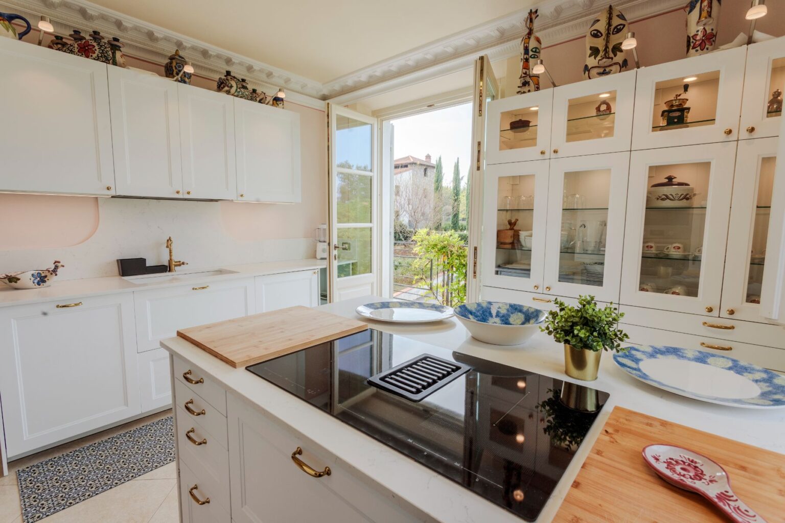 Modern kitchen interior featuring white cabinetry, a sleek stovetop, and large windows that provide natural light and a view of the garden. The design includes decorative dishes, a wooden cutting board, and potted greenery, creating a bright and inviting cooking space. | Only on SopranoVillas