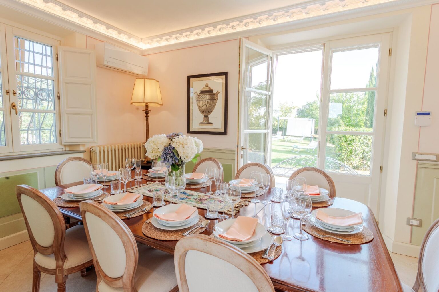 Elegant dining room featuring a beautifully set wooden table with fine china, glassware, and floral centerpiece. Sunlight streams through open French doors, showcasing a lush garden view. Classic decor elements include a lamp and framed art, creating a warm and inviting atmosphere. | Only on SopranoVillas