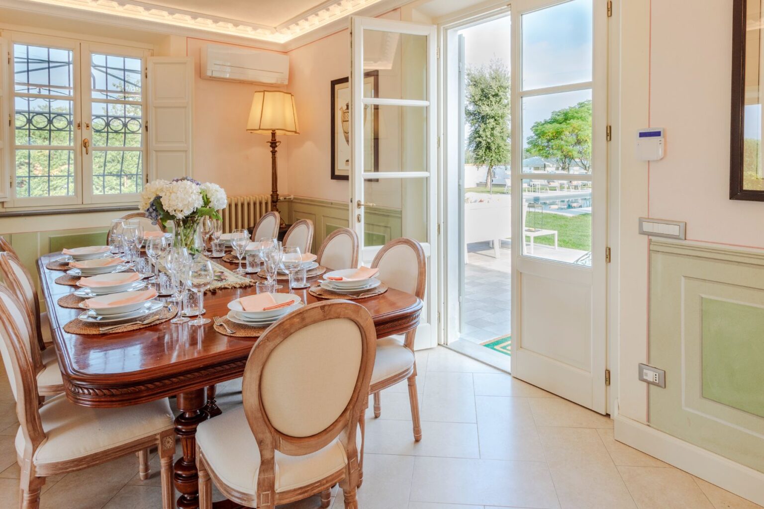 Elegant dining room featuring a large wooden table set for a meal, with fine glassware and plates. The room is illuminated by natural light from open doors leading to an outdoor area with a pool and greenery. Stylish decor includes a lamp and framed artwork on the walls, creating a warm and inviting atmosphere. | Only on SopranoVillas
