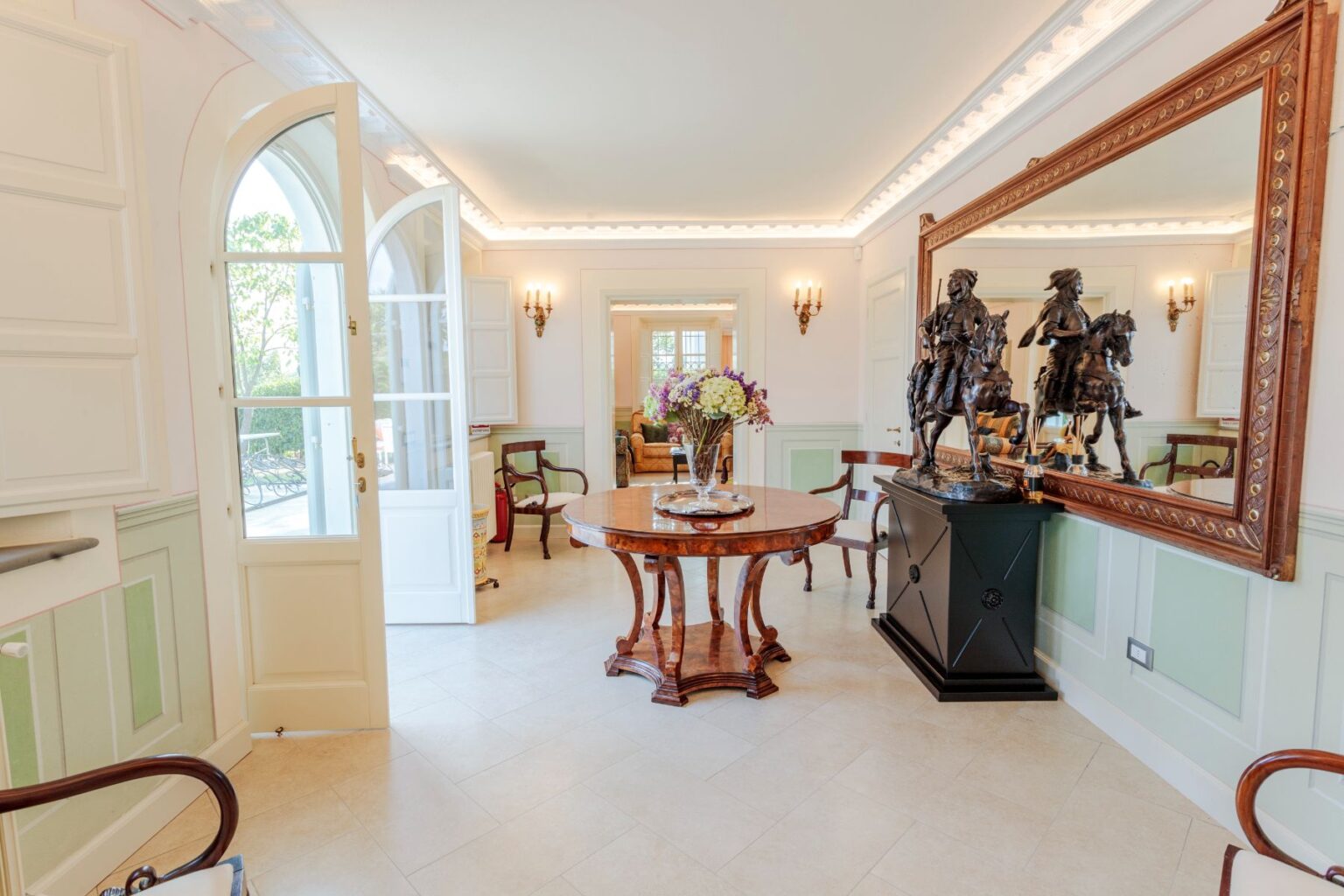 Elegant entryway featuring a round wooden table with a floral arrangement, ornate mirror, and decorative sculptures. Brightly lit with natural light from the open doors, the room showcases classic furnishings and soft pastel wall colors. Ideal for showcasing luxury interior design. | Only on SopranoVillas