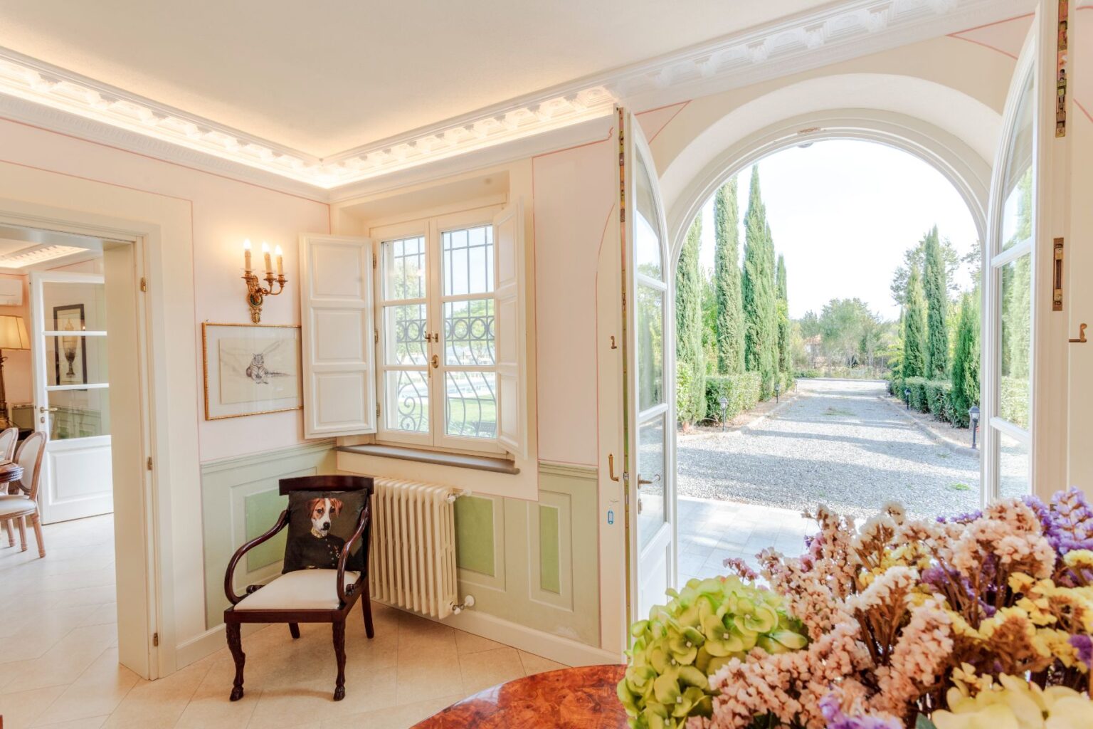 Interior view of a stylishly decorated room featuring a wooden chair with a dog-themed cushion, elegant wall sconces, and large windows that open to a landscaped garden with cypress trees. A floral arrangement is visible in the foreground, enhancing the inviting atmosphere of the space. | Only on SopranoVillas