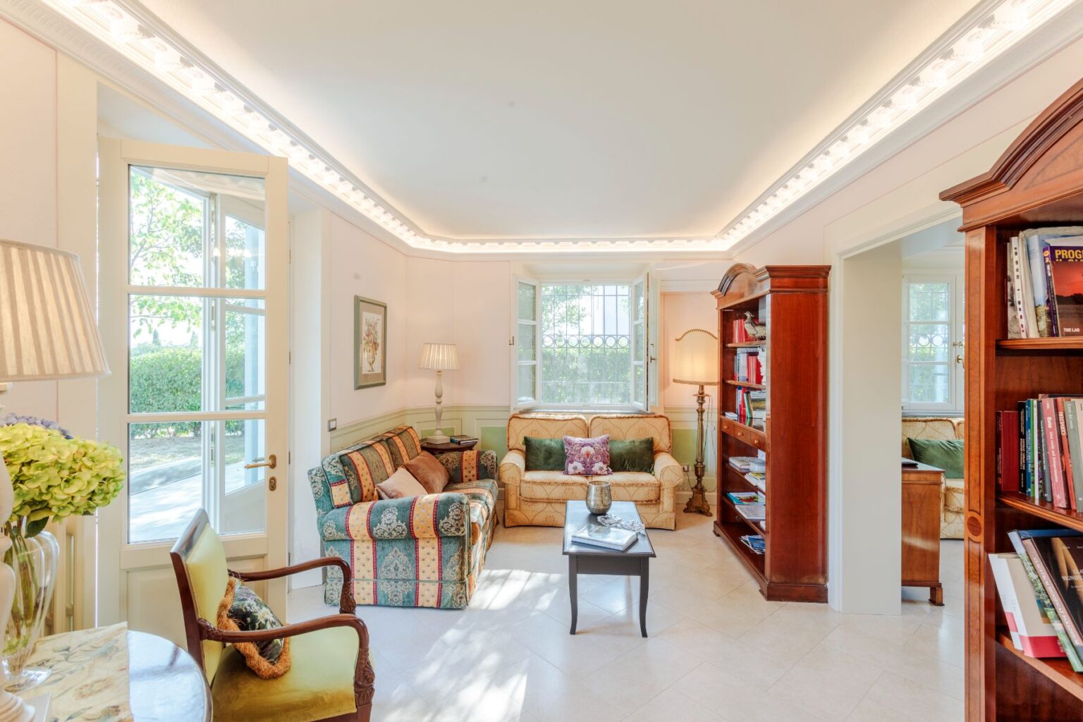 Bright and inviting living room featuring comfortable seating, elegant decor, a wooden bookshelf filled with books, and large windows that let in natural light, creating a warm and cozy atmosphere. | Only on SopranoVillas