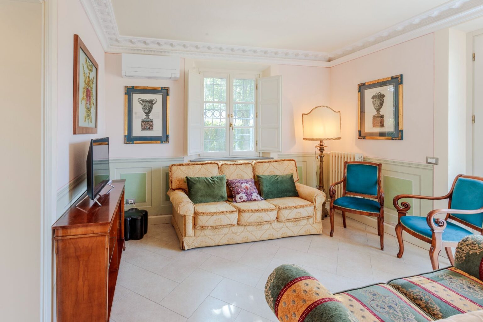 Cozy living room featuring a cream-colored sofa with green and patterned cushions, a wooden TV stand, and decorative wall art. The space is illuminated by a standing lamp and has large windows with white shutters, complemented by elegant blue chairs and a classic radiator. | Only on SopranoVillas