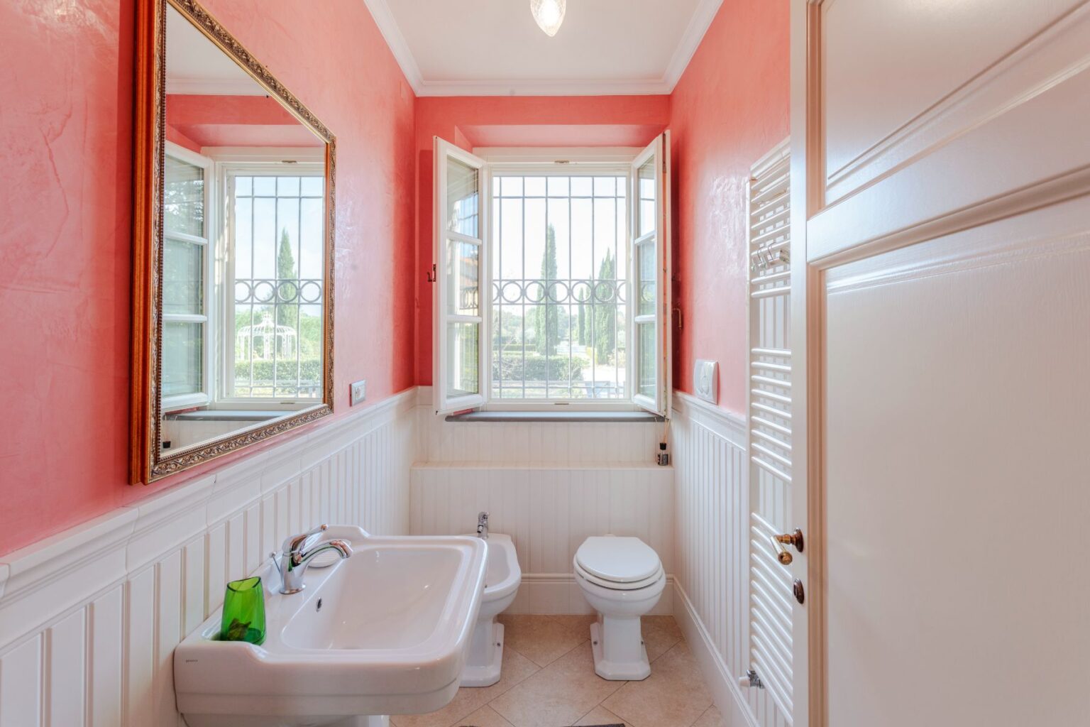 Bright and airy bathroom featuring a pink textured wall, large mirror, white fixtures including a sink and toilet, and a window with a view of greenery. The room is accented with decorative molding and a towel radiator, creating a stylish and inviting atmosphere. | Only on SopranoVillas