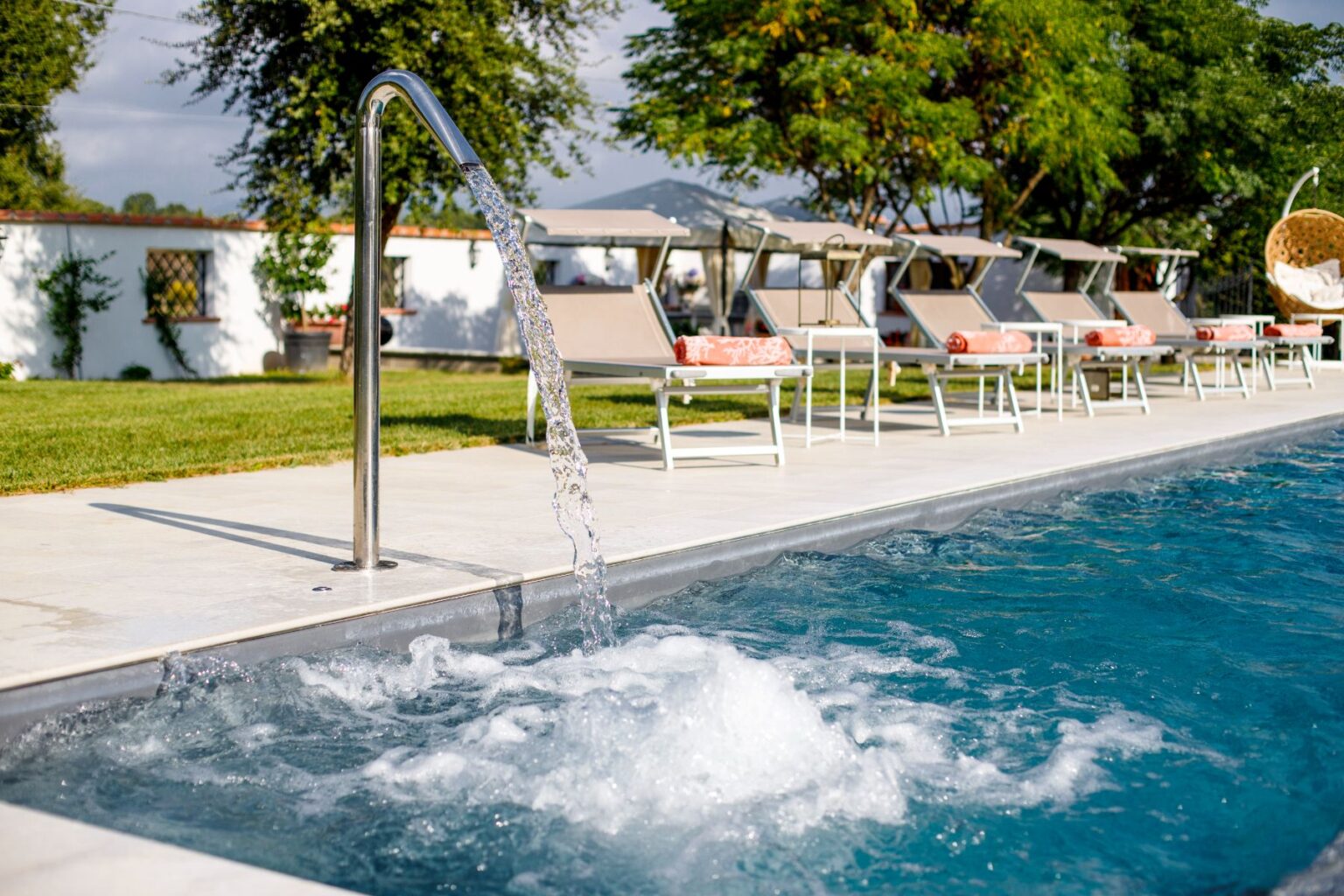 Modern swimming pool with a stainless steel water feature, bubbling water, and lounge chairs in a serene outdoor setting surrounded by greenery and a white wall. | Only on SopranoVillas