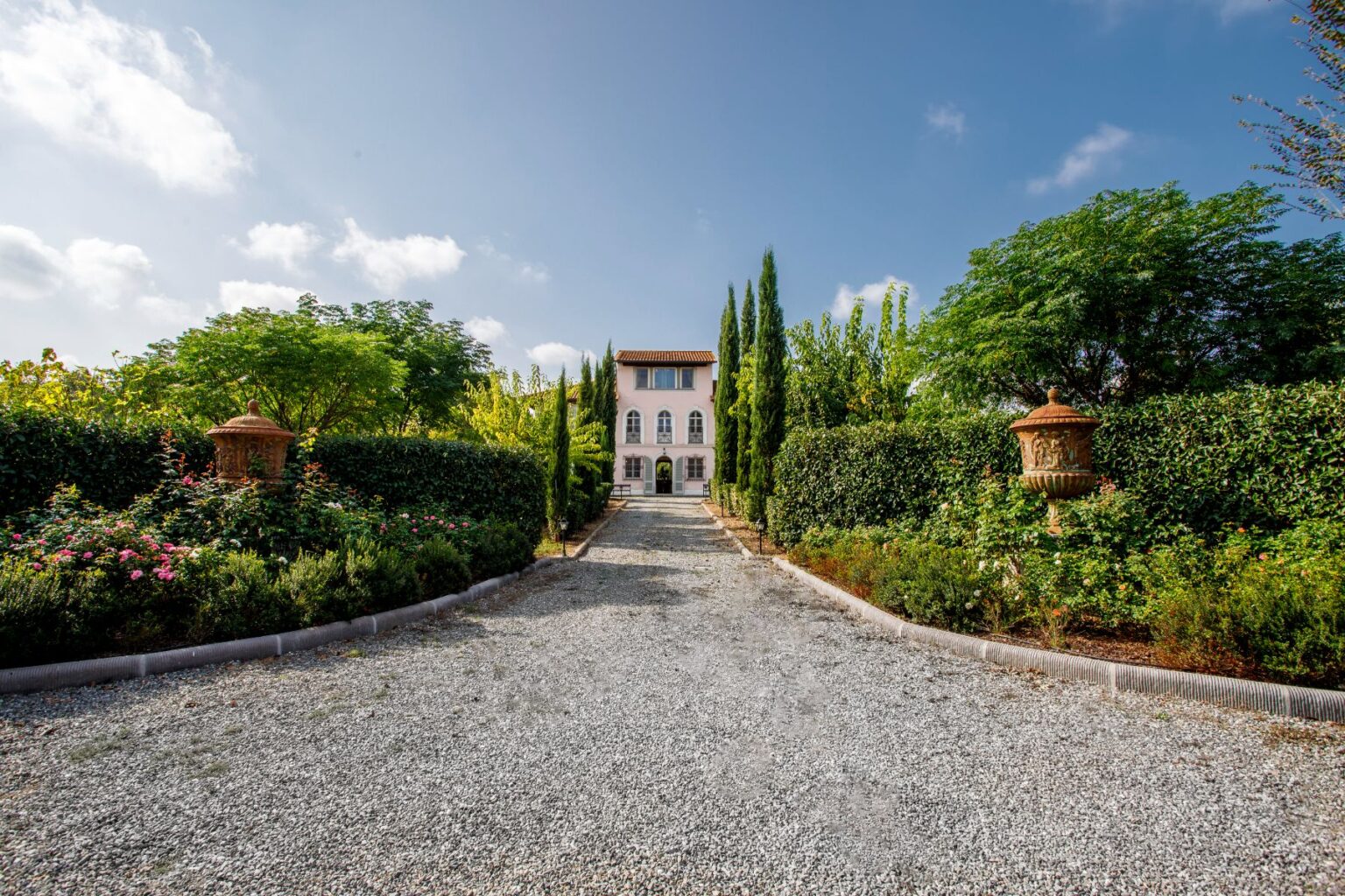 Elegant villa surrounded by lush gardens and neatly trimmed hedges, featuring a gravel pathway leading to the entrance. The scene is set against a clear blue sky, highlighting the beauty of the landscape. Perfect for showcasing luxury real estate or travel destinations. | Only on SopranoVillas