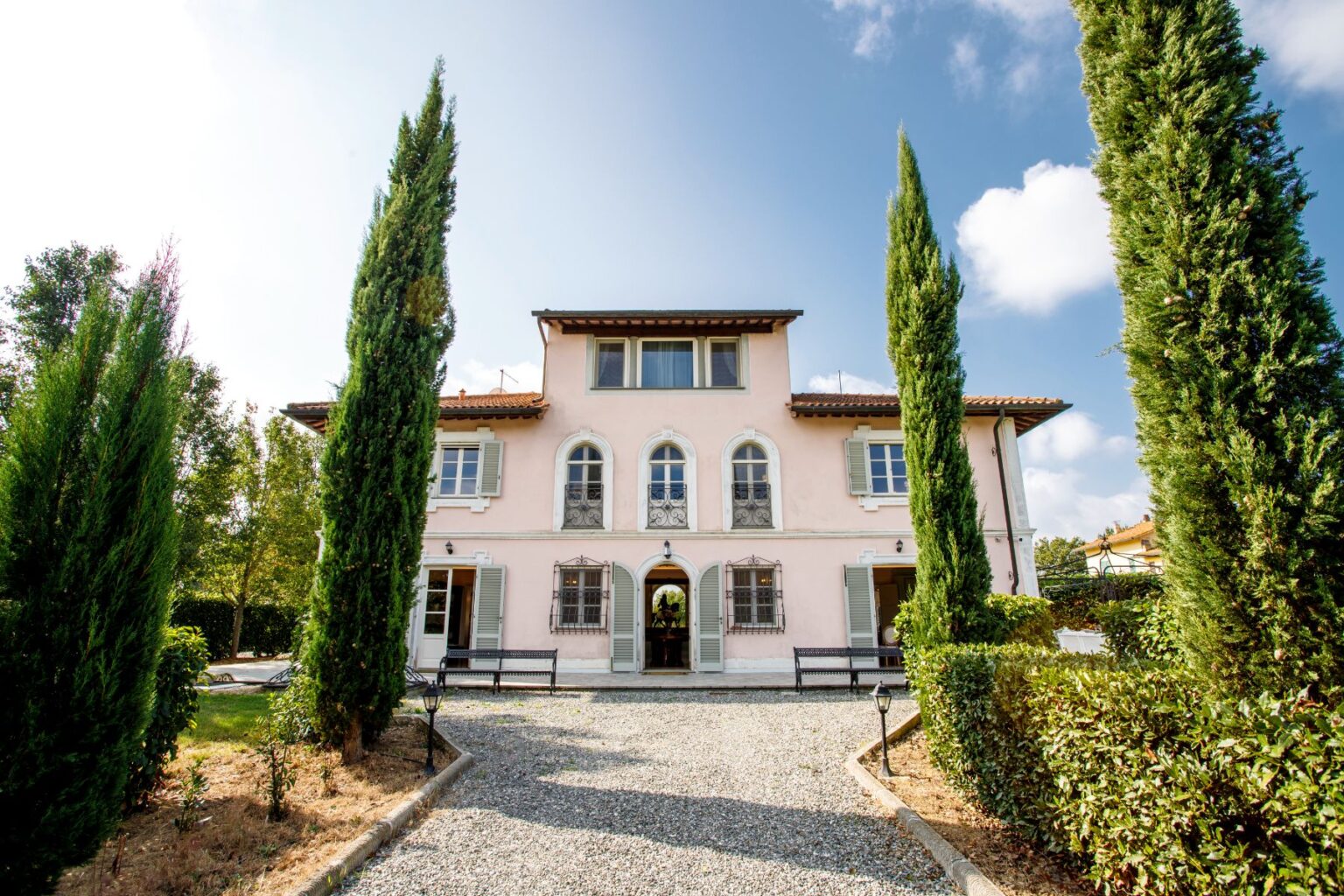Elegant pink villa surrounded by tall cypress trees, featuring a gravel pathway leading to the entrance, framed by lush greenery and a clear blue sky. Ideal for events or luxury getaways. | Only on SopranoVillas
