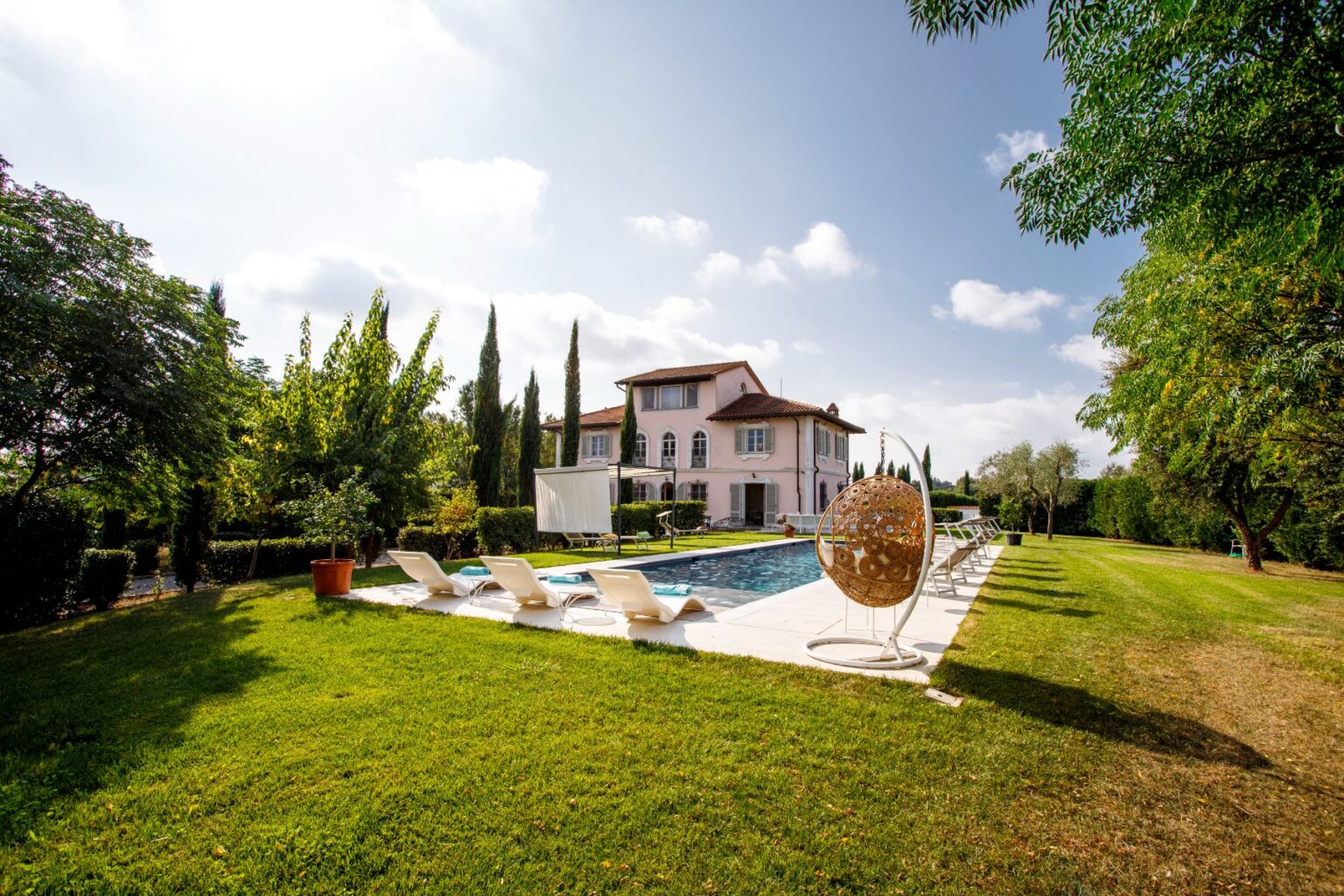 Luxury villa with a swimming pool surrounded by lush greenery, sun loungers, and a stylish hanging chair, set against a clear blue sky. Ideal for relaxation and outdoor gatherings. | Only on SopranoVillas