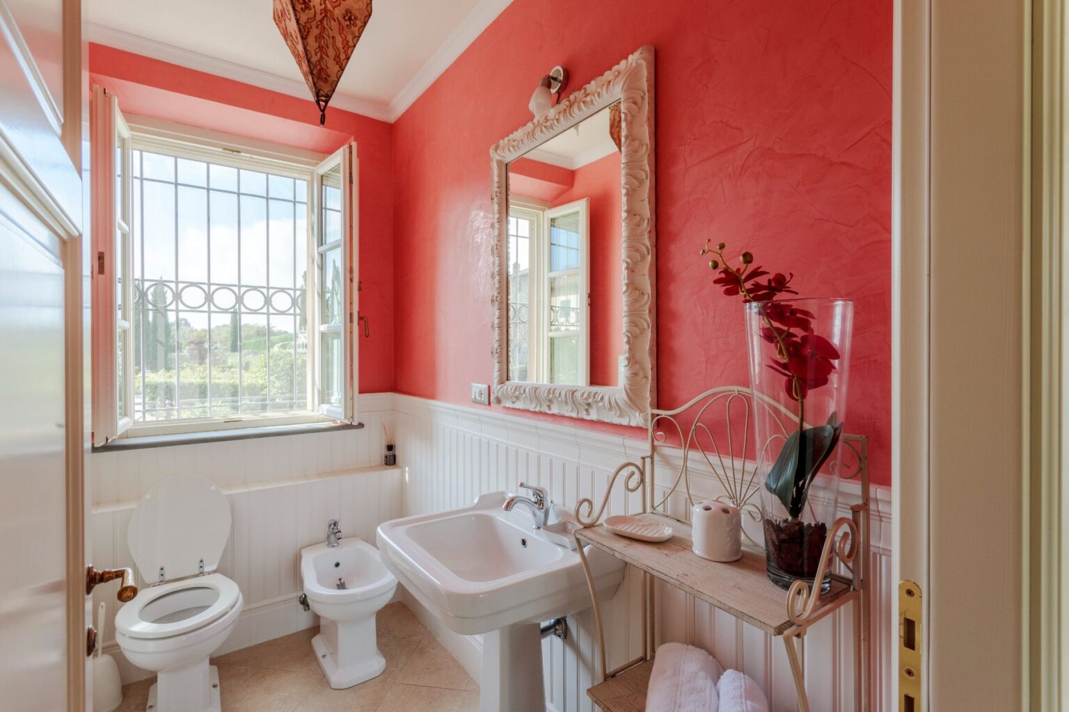 Bright and stylish bathroom featuring coral walls, a large decorative mirror, a window with a scenic view, and elegant fixtures including a toilet and bidet. The space is accented with a floral arrangement and neatly organized towels on a decorative shelf. | Only on SopranoVillas
