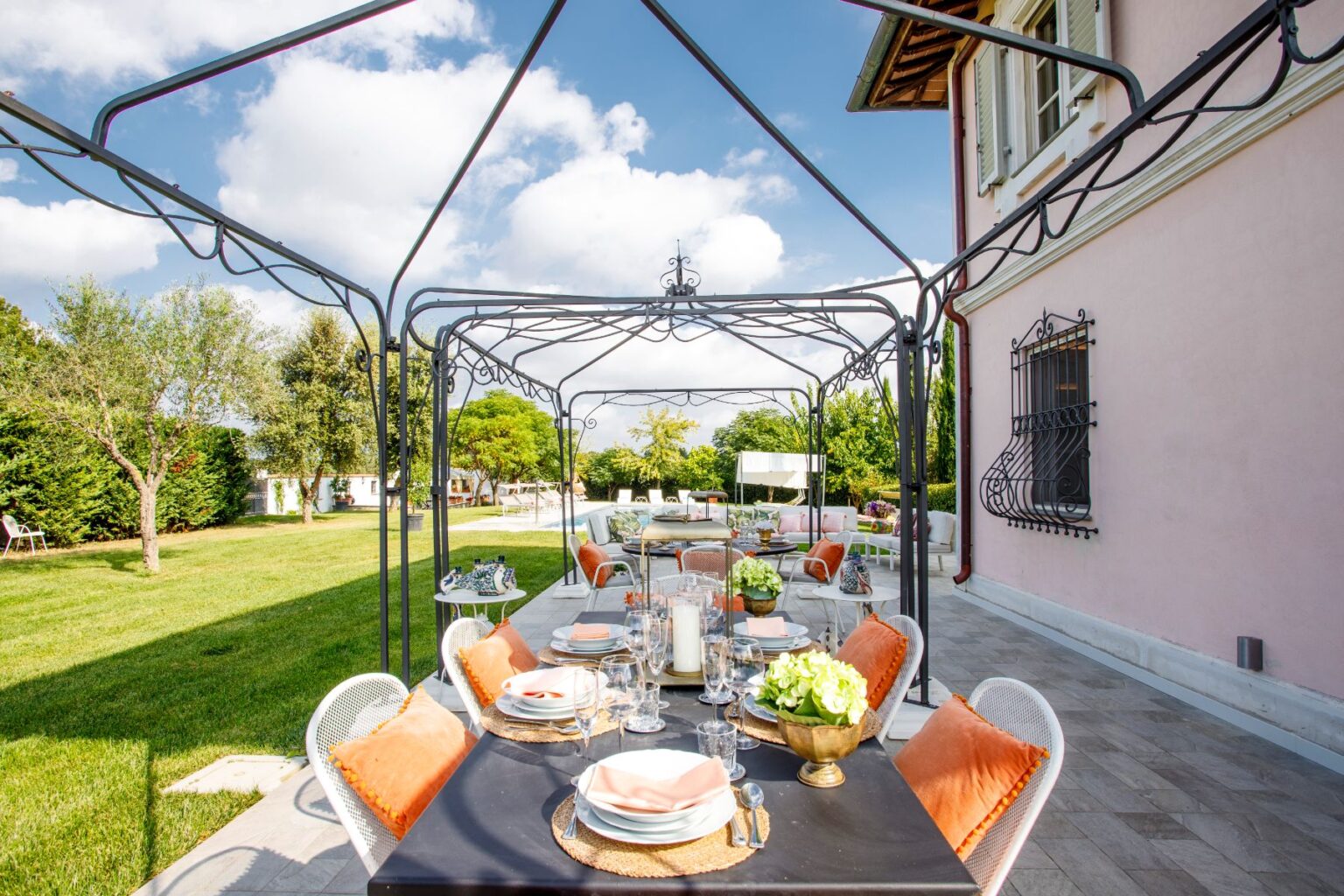 Outdoor dining area with a stylish black table set for a meal, featuring elegant dishware and floral centerpieces, under a decorative canopy. Lush green lawn and trees provide a serene backdrop, enhancing the inviting atmosphere of the space. | Only on SopranoVillas