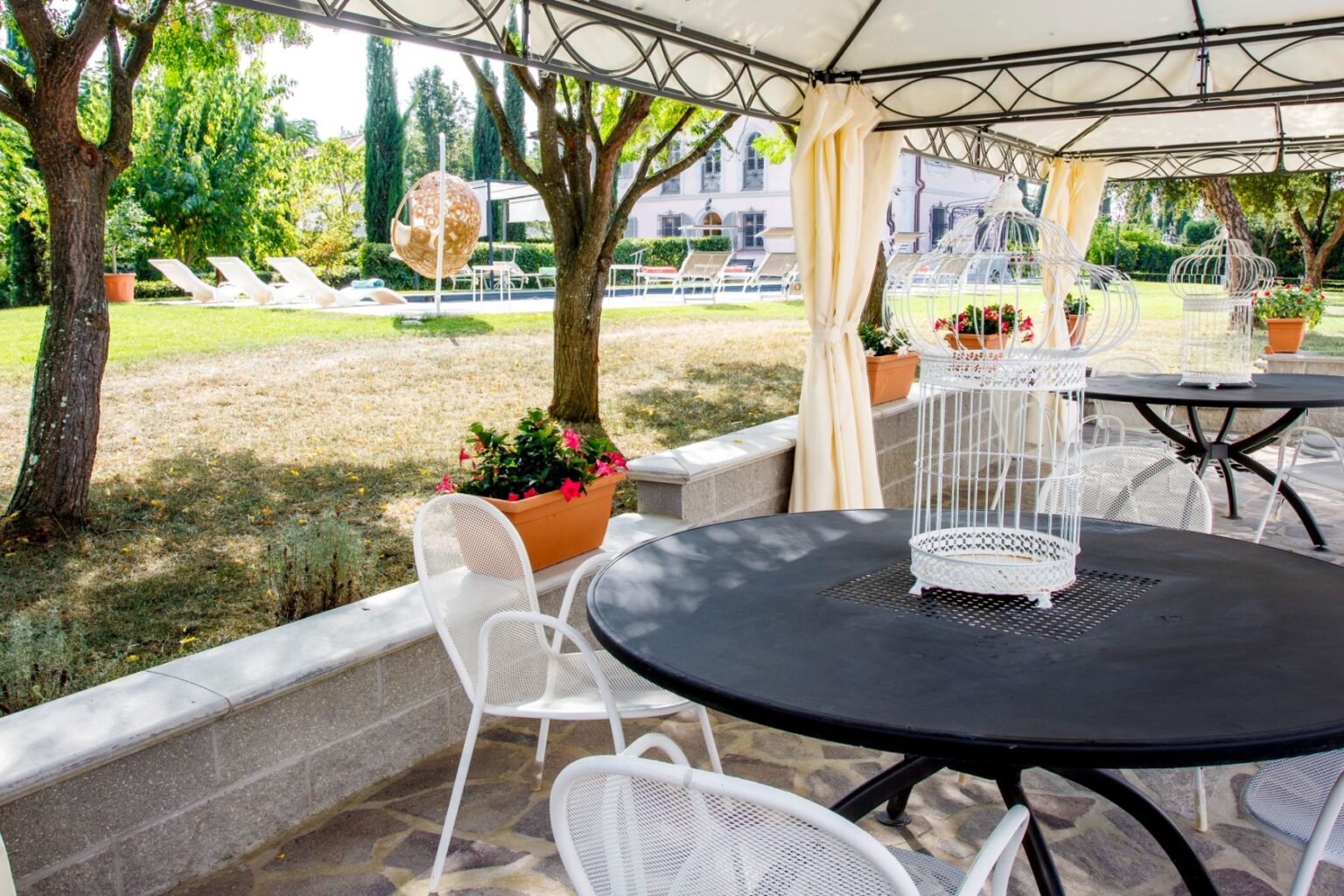 Outdoor seating area with black round tables and white chairs under a canopy, featuring decorative birdcages and potted flowers. In the background, a green lawn with sun loungers and a unique hanging chair creates a relaxing atmosphere. Perfect for enjoying sunny days in a tranquil garden setting. | Only on SopranoVillas