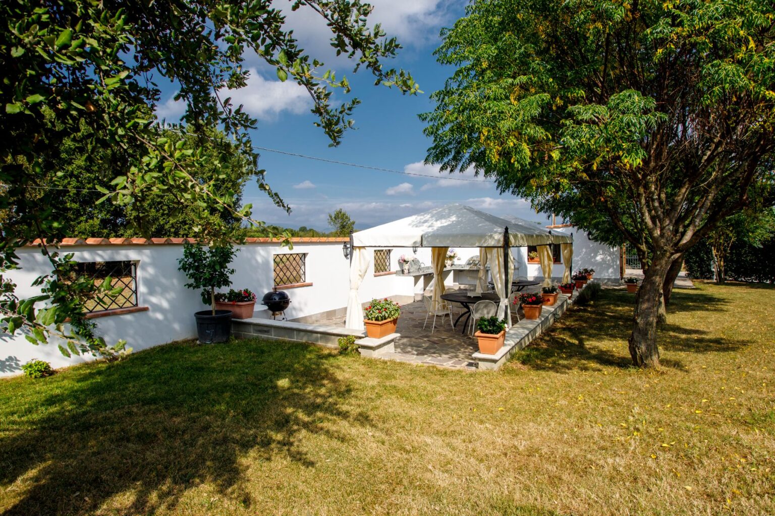 Cozy outdoor patio with a white gazebo, surrounded by lush greenery and potted plants, ideal for relaxation or gatherings. The scene features a well-maintained lawn and a barbecue grill, set against a clear blue sky. | Only on SopranoVillas