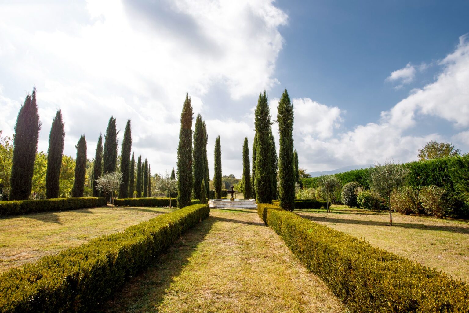 Lush garden pathway lined with tall cypress trees, bordered by neatly trimmed hedges, leading to a central fountain under a partly cloudy sky. Ideal for nature lovers and garden enthusiasts. | Only on SopranoVillas