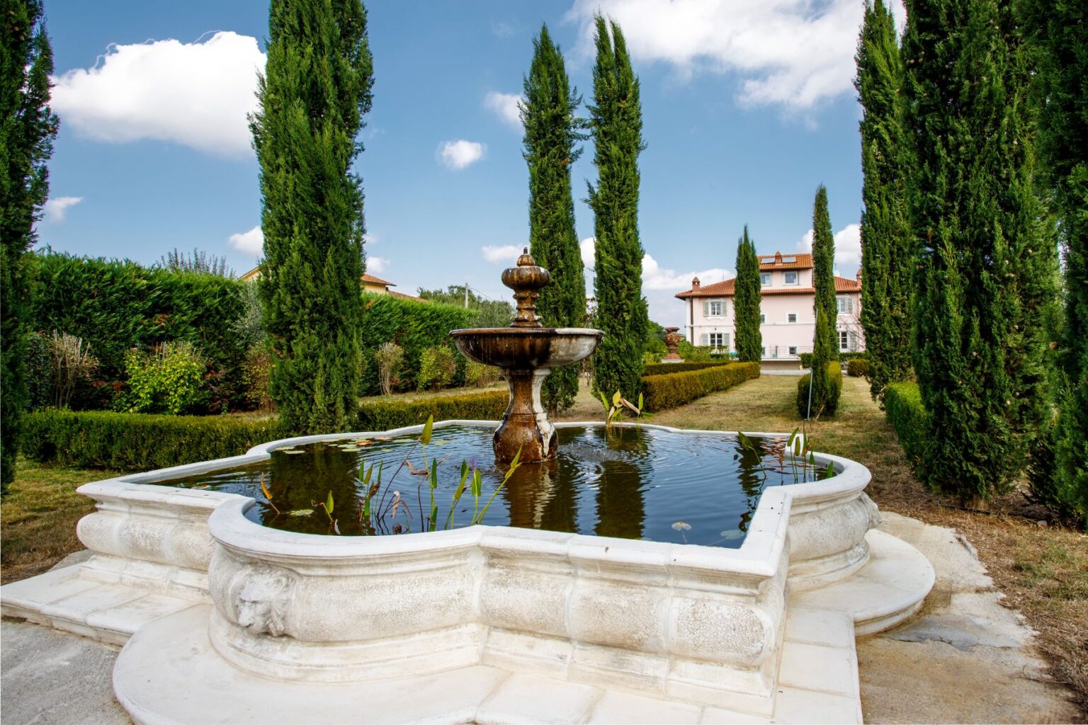Beautiful garden scene featuring a classic stone fountain surrounded by lush greenery and tall cypress trees, with a serene pond reflecting the sky and a charming pink villa in the background. Ideal for showcasing outdoor landscaping and architecture. | Only on SopranoVillas