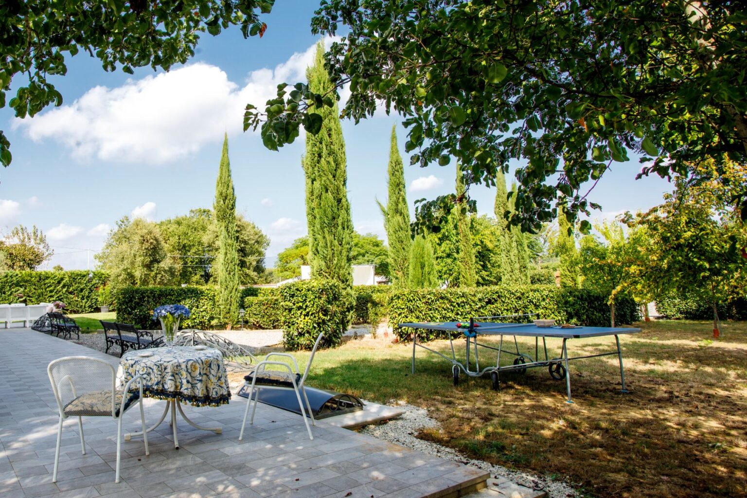 A sunny garden scene featuring a table with a floral tablecloth and chairs, surrounded by lush greenery and tall cypress trees. A trampoline is visible in the background, creating a perfect outdoor space for relaxation and recreation. | Only on SopranoVillas
