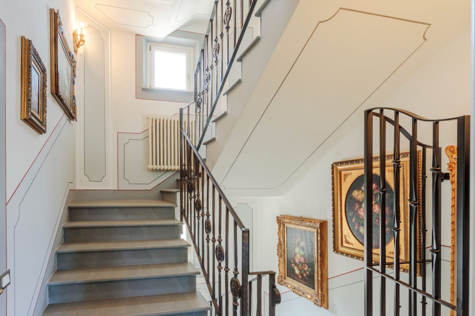 Elegant staircase with decorative wall art, featuring vintage paintings in ornate frames, complemented by a stylish wrought iron railing and a modern radiator. Natural light streams in from a nearby window, enhancing the sophisticated ambiance of the space. | Only on SopranoVillas