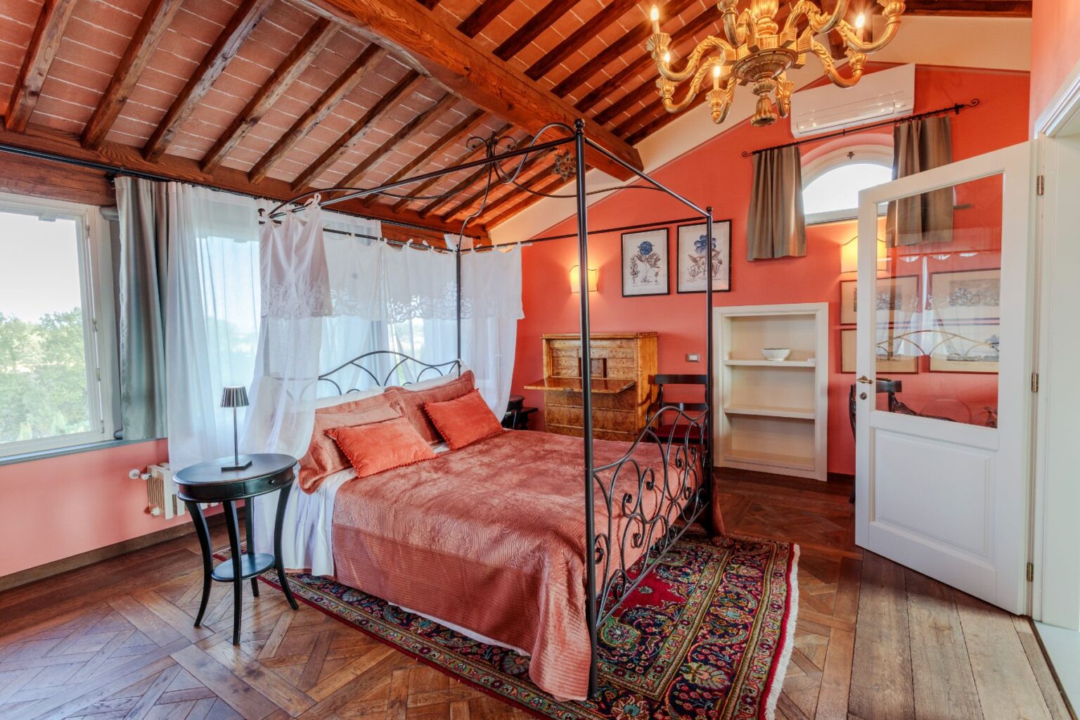 Cozy bedroom featuring a four-poster bed with decorative pillows, warm peach walls, wooden ceiling beams, and elegant chandelier. The room includes a small side table, a bookshelf, and large windows that offer natural light and a view of greenery. Ideal for relaxation and comfort. | Only on SopranoVillas