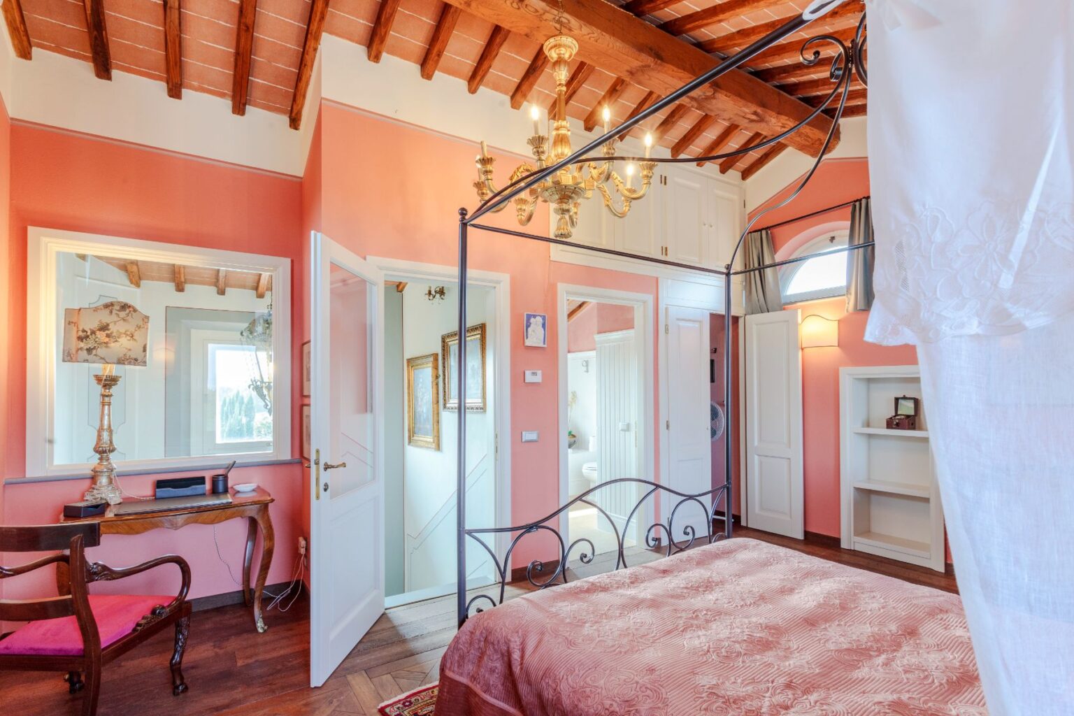 Cozy bedroom featuring a four-poster bed with a pink patterned cover, elegant wooden ceiling beams, and warm peach walls. The room includes a vintage wooden chair, a decorative lamp, and an open door leading to a bathroom. Natural light floods in through large windows, enhancing the inviting atmosphere. | Only on SopranoVillas