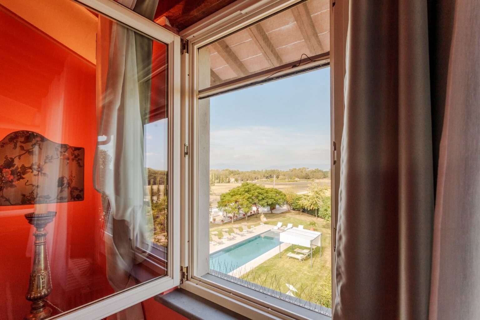 View from a window showcasing a serene outdoor scene with a swimming pool, lounge chairs, and lush greenery, framed by elegant curtains and a decorative lamp in a warm-toned interior. | Only on SopranoVillas