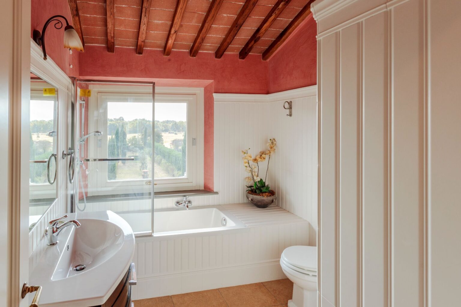 Modern bathroom interior featuring a glass shower, freestanding bathtub, and elegant fixtures. Natural light streams through the large window, highlighting the warm terracotta ceiling and stylish decor, including decorative plants. | Only on SopranoVillas