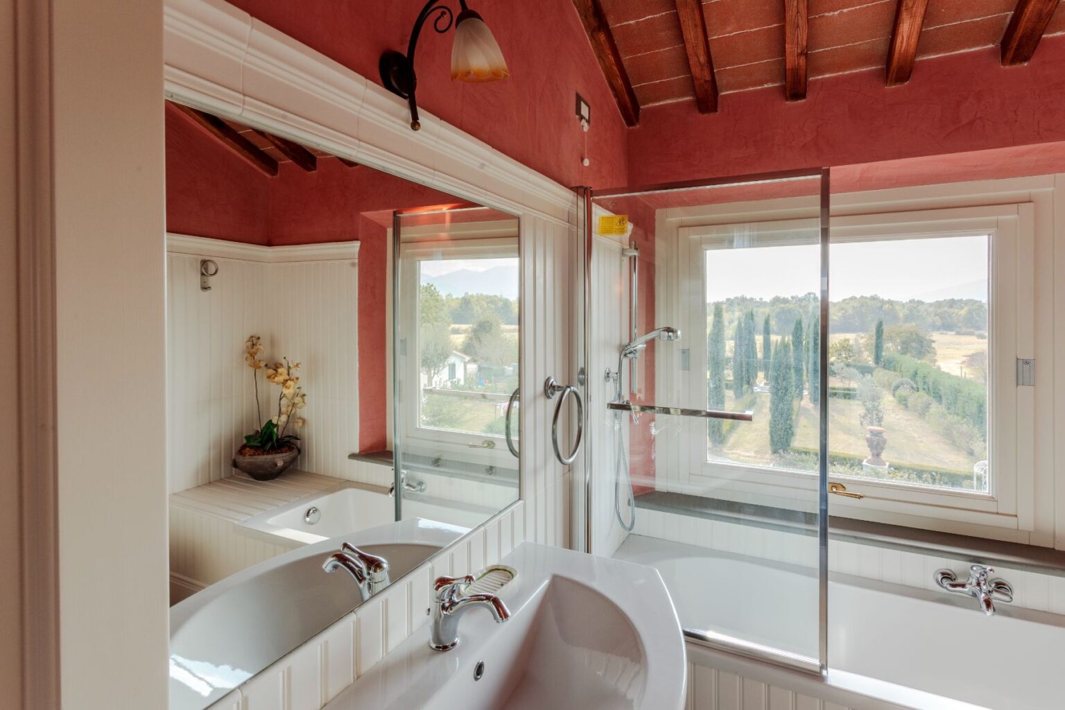 Modern bathroom with a large soaking tub, glass shower enclosure, and scenic views of the countryside through large windows, featuring rustic wooden beams and warm pink walls. | Only on SopranoVillas