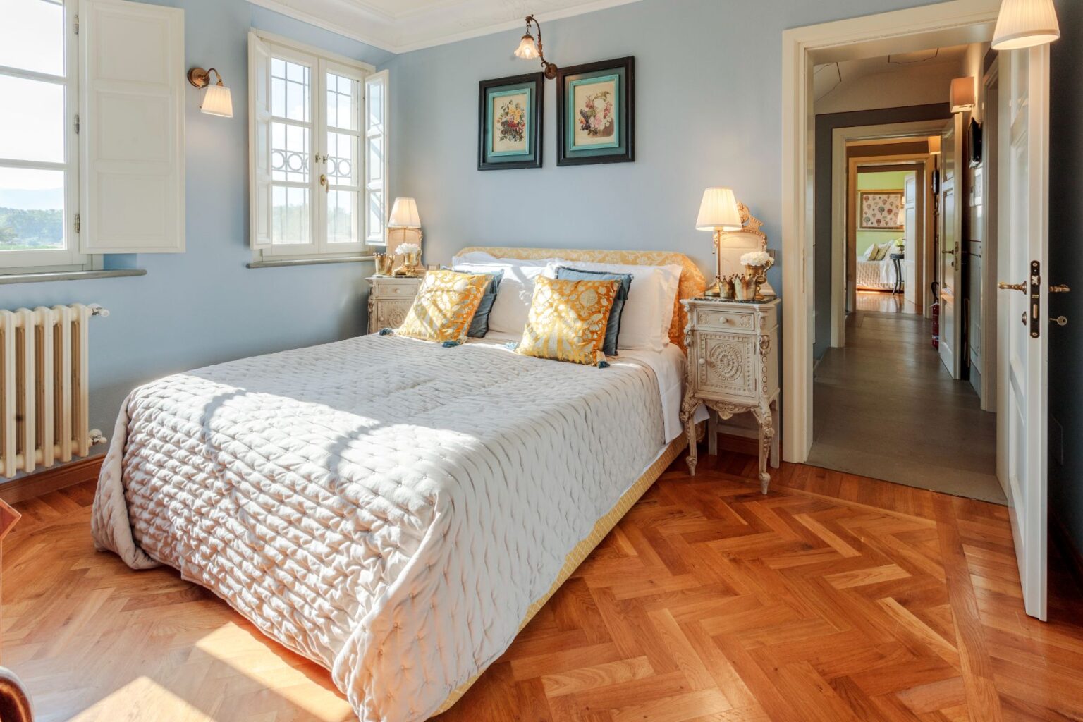 Bright and inviting bedroom featuring a cozy queen-sized bed with decorative pillows, elegant bedside tables, and large windows allowing natural light. The room showcases a soft blue wall color, hardwood flooring, and tasteful artwork, creating a serene atmosphere. | Only on SopranoVillas