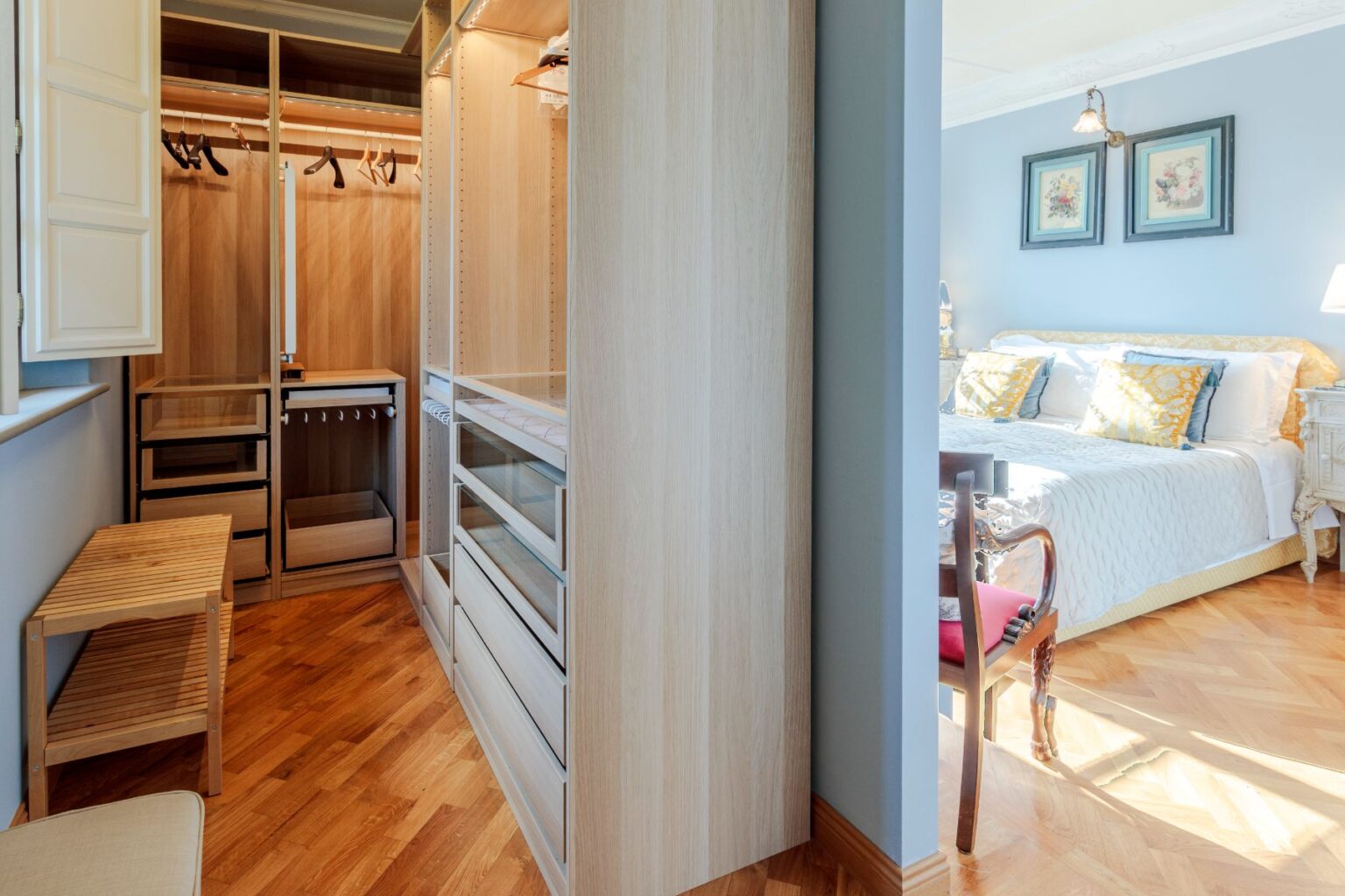 Modern bedroom featuring a stylish wooden wardrobe with open shelving, drawers, and hanging space, complemented by a cozy bed adorned with decorative pillows and elegant artwork on the walls. Natural light enhances the warm tones of the hardwood floor. | Only on SopranoVillas