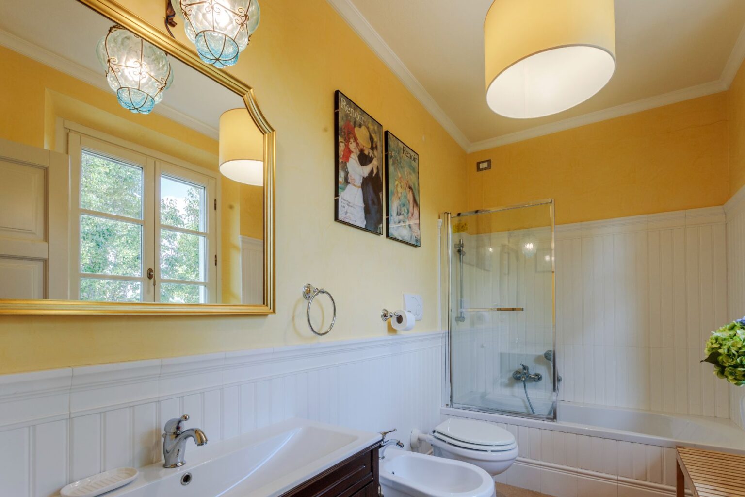 Bright and inviting bathroom featuring yellow walls, elegant glass light fixtures, and a large mirror. The space includes a bathtub with a glass shower enclosure, a modern toilet, and stylish artwork by Renoir. Natural light streams in through a window, enhancing the warm atmosphere. | Only on SopranoVillas